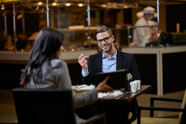 Glad man with short beard wearing glasses and looking with interest at picture on tablet, presented by business partner