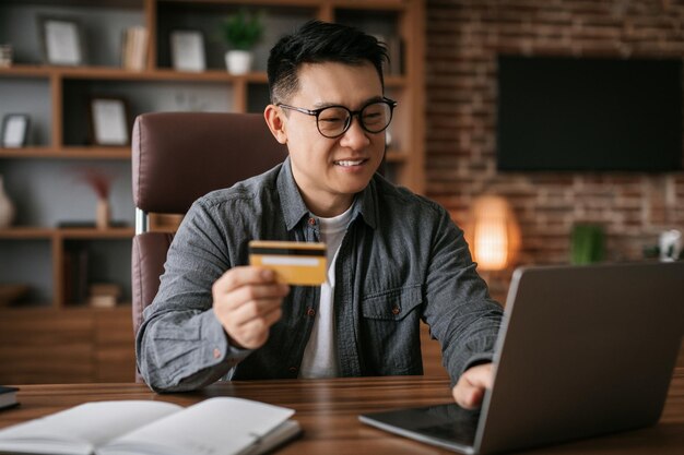 Glad mature asian man in glasses typing on laptop showing credit card at table in home office interior