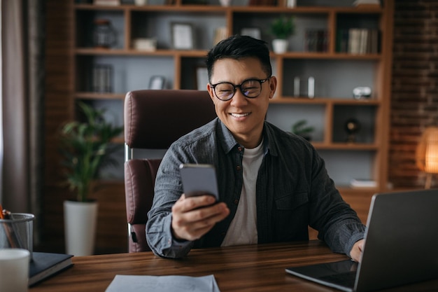 Il maschio coreano maturo felice con gli occhiali legge il messaggio sul lavoro dello smartphone sul laptop nell'interno dell'ufficio domestico