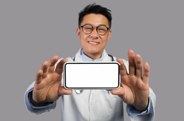흰 코트와 안경을 쓴 다행스러운 중년 아시아 남자 치료사는 빈 화면으로 스마트폰을 보여준다