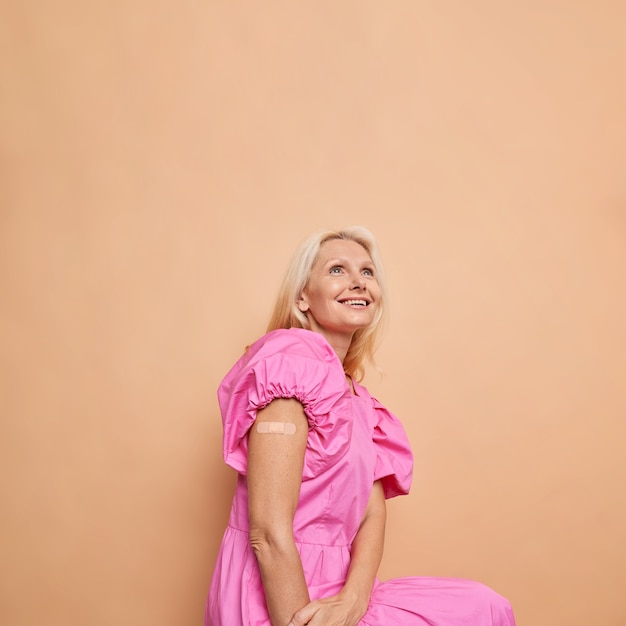 Glad middle aged blonde woman shows shoulder with band aid after getting vaccine involved in immunization against coronavirus beige studio wall.