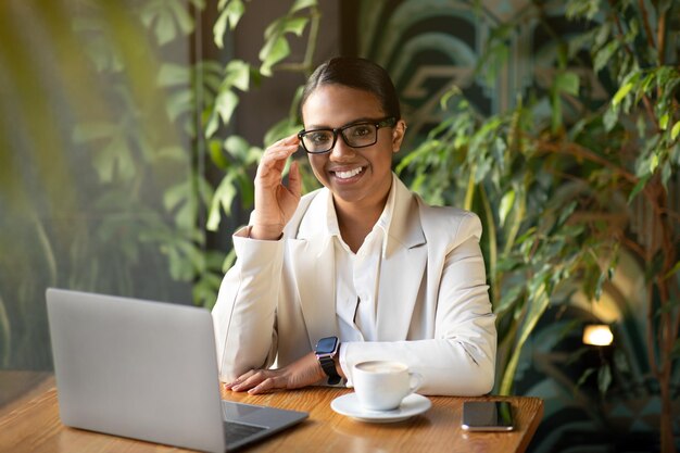 Glad millennial african american businesswoman in white suit and glasses work with laptop in cafe
