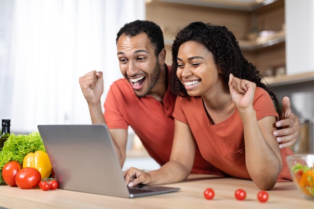 Glad millennial african american family at table with organic vegetables uses laptop make victory gesture