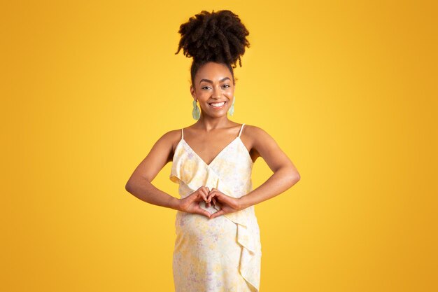 Glad millennial african american lady with belly making heart sign with hands isolated on yellow