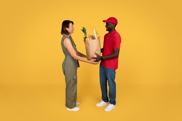 Glad millennial african american man courier in uniform give paper bag with food to adult woman