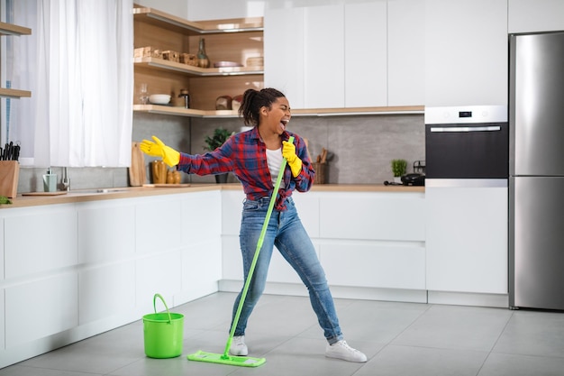 Glad millennial black woman housewife in rubber gloves with mop washes floor sings song and dances