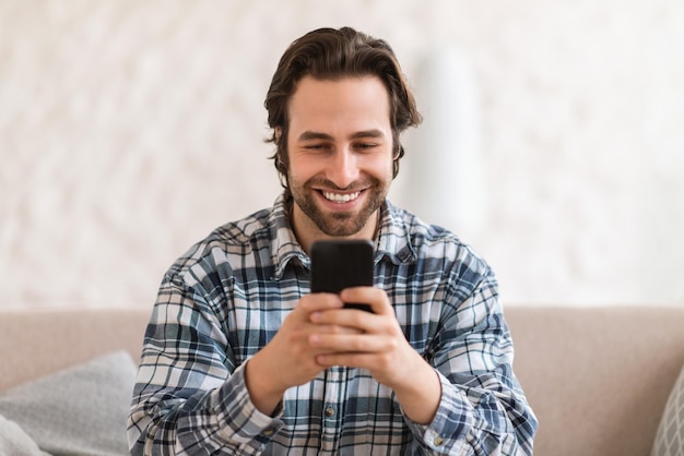 Glad millennial caucasian man in domestic clothes with stubble on sofa chatting on smartphone