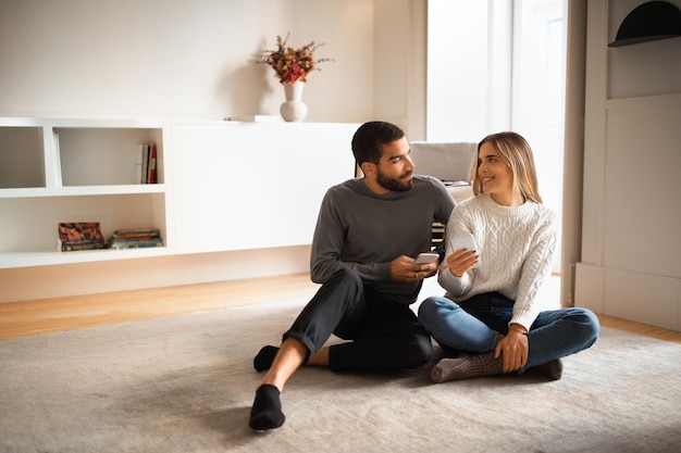 Glad millennial european woman arab male talk typing on smartphone reading message sitting on floor