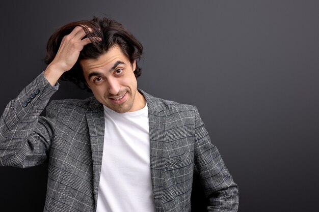 Glad millennial guy in beautiful wear suit looking at camera touching hair, isolated on black background