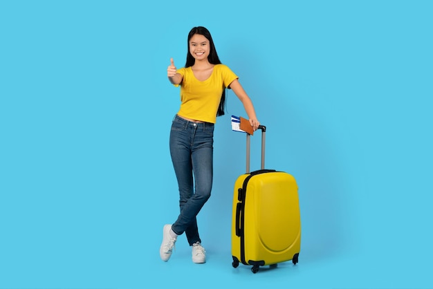 Glad millennial korean lady student in yellow tshirt with suitcase and tickets show thumb up