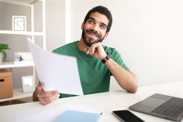 Glad pensive young arab businessman at workplace with laptop works with documents look at free space