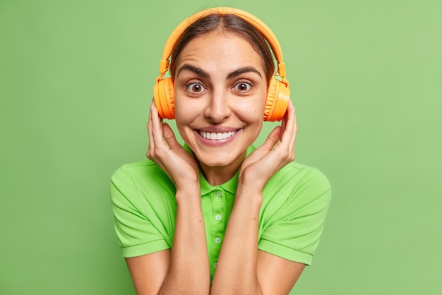 Glad pleasant looking woman with dark hair enjoys listening
music via wireless headphones smiles toothily wears casual t shirt
isolated over vivid green background listens favorite
composition