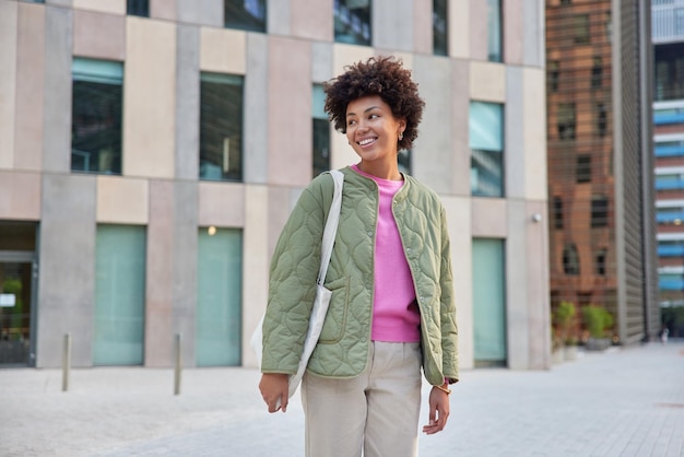 Glad positive woman in jacket and trousers carries bag looks away happily poses against modern urban building concentrated away enjoys spare time during weekend People and lifestyle concept