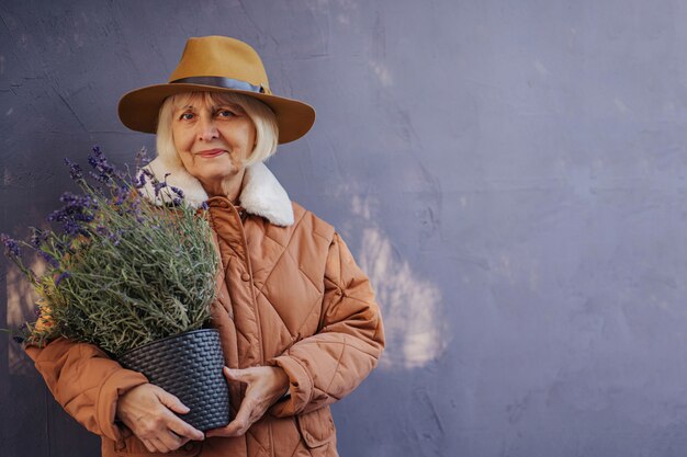 Glad senior female in stylish outerwear and hat smiling for camera and carrying potted lavender