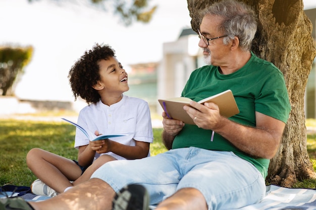 Glad senior man with beard and black little boy sit on plaid read a book relax in park