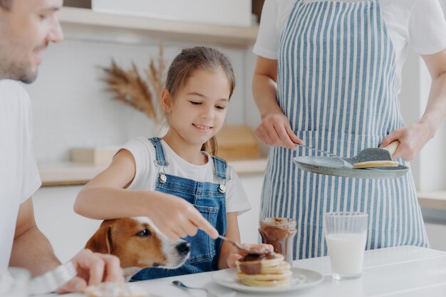 Glad small girl enjoys eating tasty dessert prepared by mum adds melted chocolate to pancakes enjoys being together and mother father and dog have delicious nutritious breakfast in kitchen