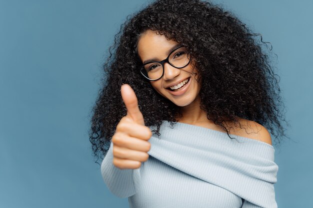 Glad smiling African American woman shows like gesture, keeps thumb up, gives approval