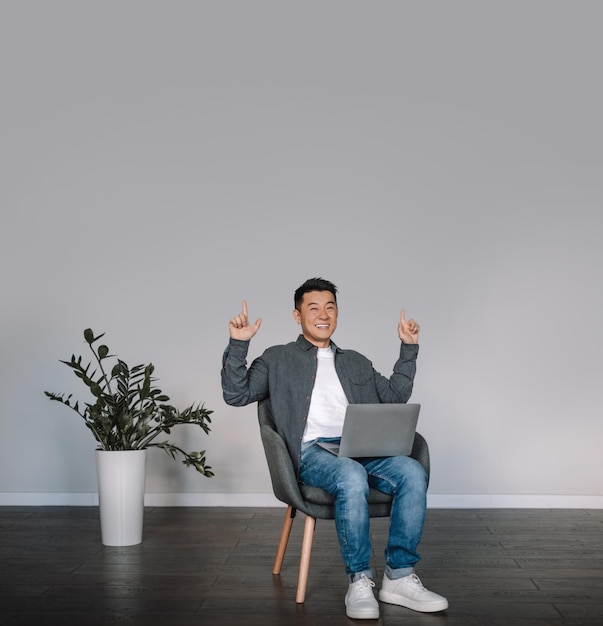 Glad smiling middle aged chinese male with laptop making victory gesture with hands sit in armchair