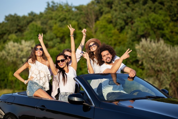 Glad stylish young dark-haired girls and guys in sunglasses smile in a black cabriolet on the road holding their hands up on a sunny day. .