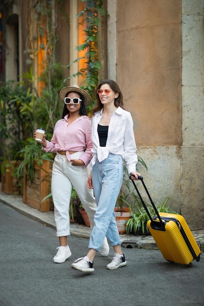 Glad stylish young millennial diverse women tourists in hat and sunglasses with suitcase and cup of