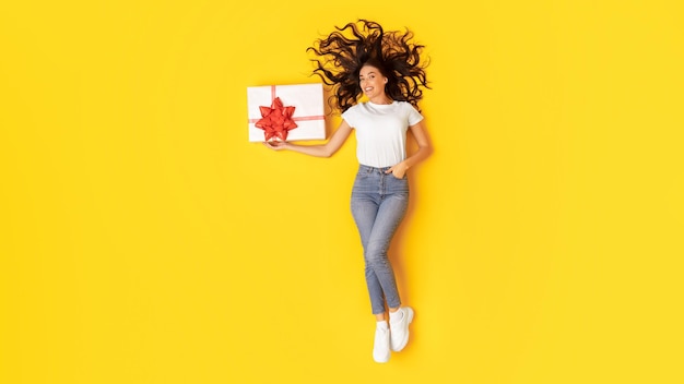 Glad woman holding wrapped present box lying over yellow background