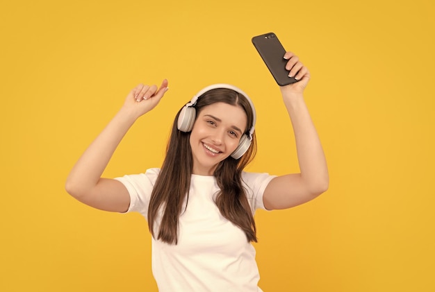 Glad woman listen music in earphones with smartphone on yellow background gadget