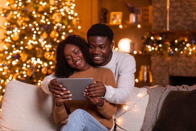 Glad young african american couple have video call on tablet watch video sit on sofa in living room