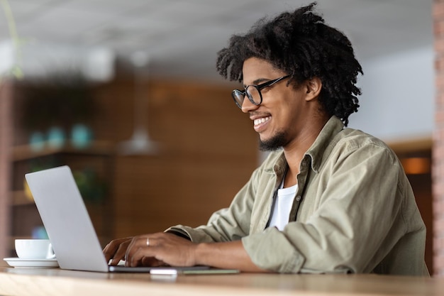 Glad young african american curly male in glasses works at laptop at table with cup in cafe interior