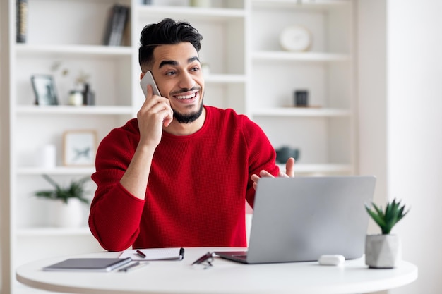 Glad young arabic male with beard speaks on phone with client at workplace with laptop in home