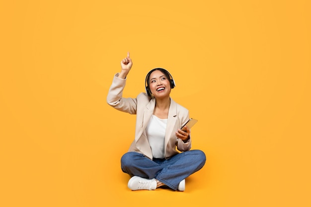 Glad young asian woman in wireless headphones with smartphone has fun singing sit on floor