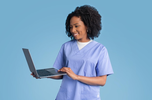 Glad young black lady doctor nurse in blue uniform uses laptop for video call