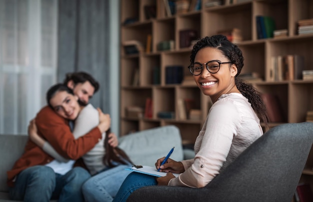 Glad young black woman doctor psychologist in glasses look at camera smiling european couple hug in