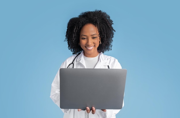 Glad young black woman doctor therapist in white coat typing on laptop has video call