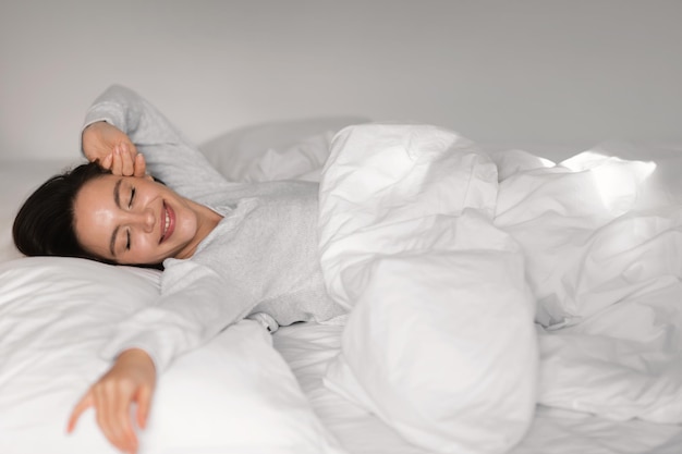 Glad young european lady sleeping on white bed with closed eyes waking up stretching body enjoying good morning