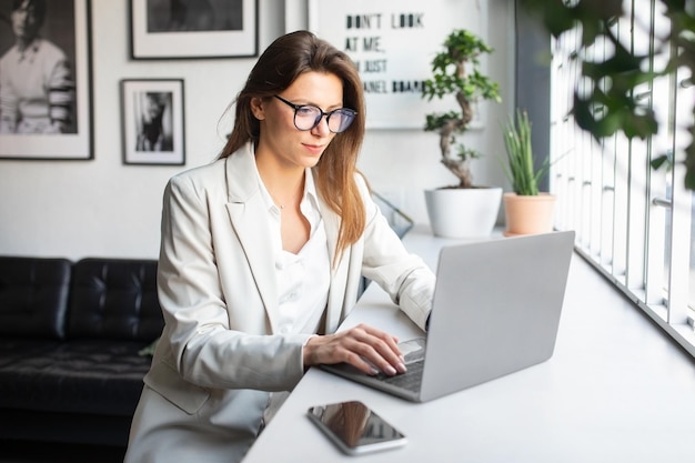 Glad young woman in suit and glasses working with laptop at table typing on computer in cozy