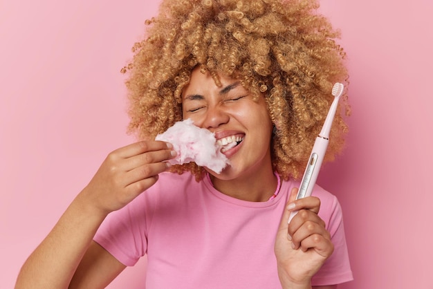 Glad young woman with curly bushy hair has sweet tooth eats candy floss going to clean teeth with electric brush dressed in casual t shirt isolated over pink background Food bad for your teeth