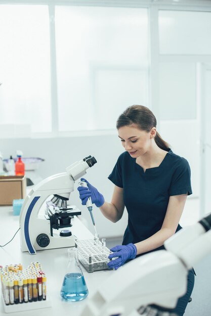 Gladsome lab scientist sitting at the table and using an automatic pipette dispenser for the liquids in test tubes