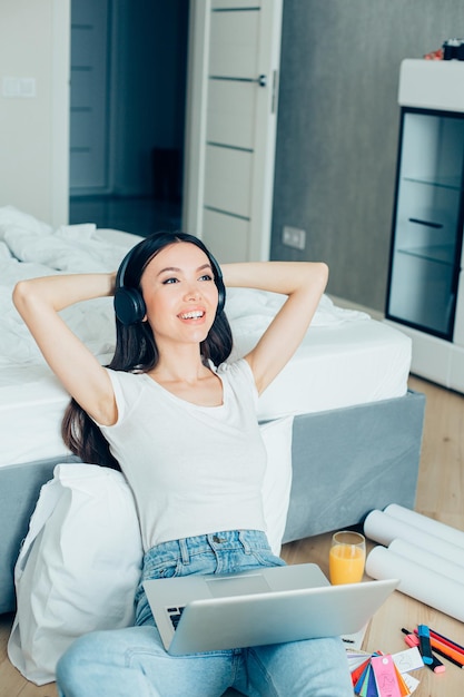 Photo gladsome lady putting hands behind her head and looking happy while sitting with a laptop and blueprints on the floor