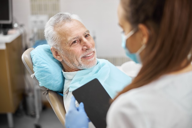 Радостный старший мужчина сидит в стоматологическом кресле и смотрит на стоматолога-эксперта, держащего планшет