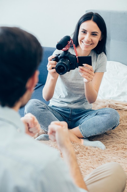 Gladsome young lady with a camera smiling and looking at the\
man in front of her and recording him on a camera