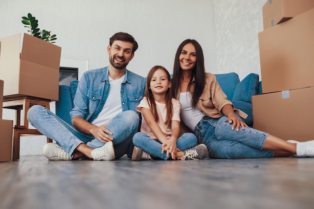 Gladsome young man with wife and daughter sitting on a floor between stacks of boxes with packed things