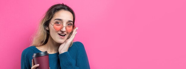 Glamor woman in glasses in a blue sweater with a drink of coffee on a pink background