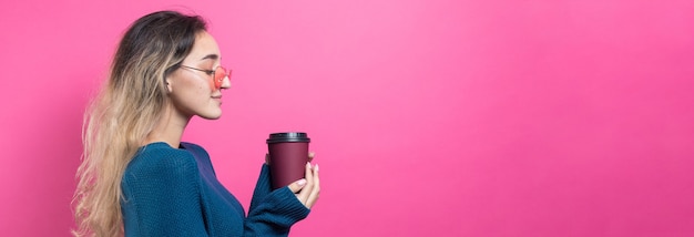 Glamor woman in glasses in a blue sweater with a drink of coffee on a pink background