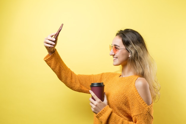 黄色の背景にコーヒーを飲みながらオレンジ色のセーターのメガネの魅力的な女性