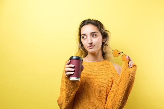 黄色の背景にコーヒーを飲みながらオレンジ色のセーターのメガネの魅力的な女性