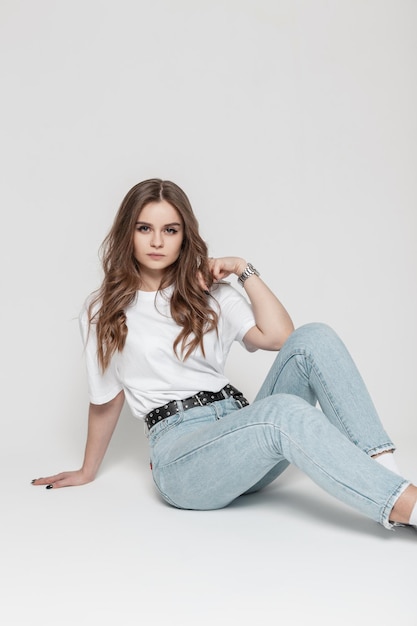 Glamorous beautiful serious girl model in casual clothes with a white tshirt and blue high waist  jeans sits in the studio