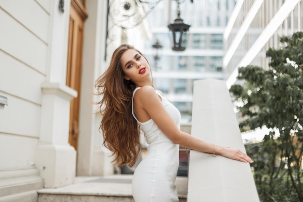 Glamorous beautiful woman model with brunet hairstyle in fashionable elegant white dress stands and poses near a white vintage building on the street