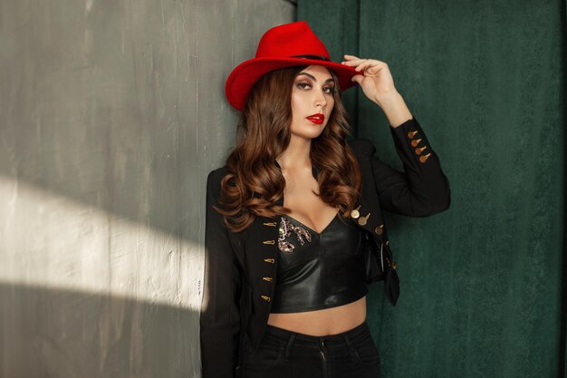 Glamorous beautiful young model woman with makeup and hairstyle in fashionable clothes with a red hat stands near a vintage wall and green curtains in the studio