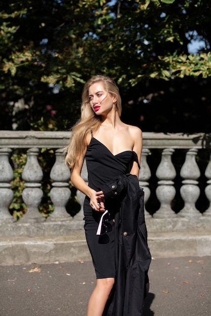 Glamorous blonde model with long hair and red lips wearing black dress, takes off her coat