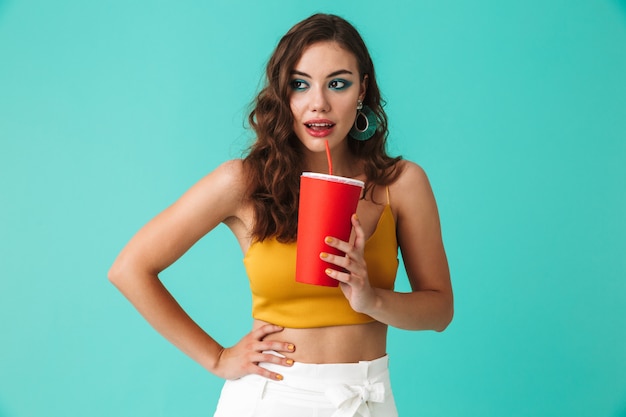 Glamorous brunette woman wearing fashion earrings drinking beverage or soda from big red paper cup and straw